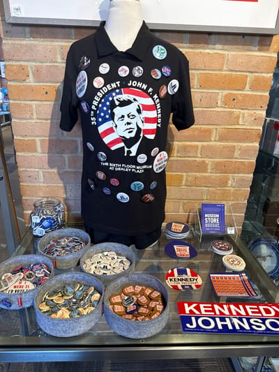 A black collared shirt, displayed on a mannequin, is adorned with various political buttons, centered around a portrait of John F. Kennedy. Buttons promoting Kennedy and Johnson are visible. Adjacent to the shirt, a glass display case holds assorted Kennedy-themed merchandise. This includes buttons organized in gray felt containers, a jar full of blue and white pins, and items like “Coffee Kennedy” mugs. A small sign for the museum store is also present, adding to the museum souvenir display.