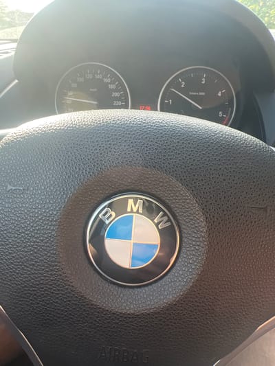 The image depicts the interior of a BMW vehicle, specifically focusing on the steering wheel and the instrument cluster, located in the city of Tsarkva, Bulgaria. The steering wheel, constructed from black textured material, prominently features the BMW logo in its center, which includes the letters "BMW" encircling a blue and white quartered motif. The word "AIRBAG" is visible at the bottom of the steering wheel's central hub, and a horn symbol is present on the left side.
The instrument cluster, partially obscured by bright sunlight, displays two primary dials. The left dial is a speedometer, calibrated in kilometers per hour (km/h), with its needle indicating approximately 50 km/h. The right dial is a tachometer, measuring revolutions per minute (1/min x 1000), with its needle positioned just below 1.5. A digital display, illuminated in red, shows the time as 17:56. Strong sunlight enters the vehicle, creating glare across the instrument panel and the upper portion of the steering wheel. Green foliage is faintly visible through the windshield in the background. No individuals are depicted in the image.
