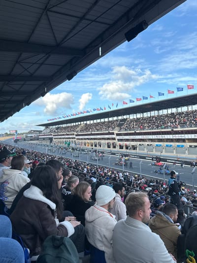 The image depicts a crowded grandstand overlooking a race track and pit lane area at the Circuit de la Sarthe in Le Mans, France. The perspective is from beneath a dark, corrugated metal roof structure, which is supported by a metal framework and has multiple black loudspeakers affixed to its underside.
In the foreground, numerous spectators are seated, primarily seen from behind, facing the track. Key individuals include a woman with long dark hair wearing a brown jacket with white sherpa lining on the collar and cuffs, a man in a white knit cap and light-colored fleece hoodie, and another man in a light grey jacket. All foreground spectators appear to be observing the activity on the track.
Beyond the immediate seating area, the asphalt race track and an adjacent pit lane are visible. Several open-wheel race cars are present on the track and in the pit lane, some stationary, while others may be in motion. At least two red and blue liveried cars are discernable on the track. Personnel, including individuals in dark uniforms, are present near the cars on the track surface. The pit lane features garage entries, some with open bays, across from the track. Advertising for "SAMSUNG Galaxy A+" is visible on trackside barriers and walls.
On the opposite side of the track, a multi-tiered grandstand is densely packed with a large number of spectators. Along the top edge of this grandstand, numerous national and sponsor flags are displayed, including several French flags. The sky above is partially cloudy, with patches of blue visible. The overall environment indicates an active motorsports event.