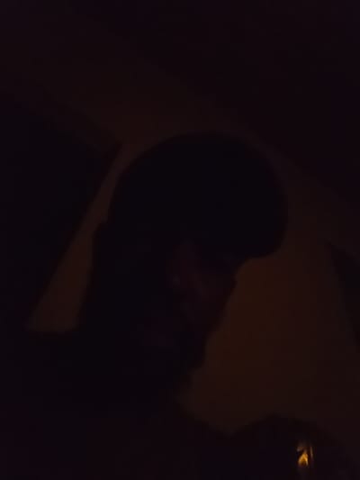 The media file shows a close-up, dimly lit shot of a person's head and shoulders, appearing to be in profile. The lighting is very low, casting the majority of the image in shadow. A warm, orangish glow is present on the right side of the frame, illuminating some of the person's features and casting a soft light on the background. The person has dark, short hair and appears to be looking downwards. The overall mood is somber and intimate due to the low lighting and close framing. It's difficult to ascertain the specific activity or event taking place, but the composition suggests a personal moment. Given the location context of Alagbado, Nigeria, this could be an indoor or outdoor setting at night. No text is visible in the image.