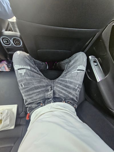 A high-angle shot captures a person's legs in the back seat of a car, with their knees bent and spread apart. They are wearing distressed grey jeans with visible rips on the thighs and a white t-shirt. The car interior is visible, featuring black leather seats, air vents on the back of the driver's seat, and a silver electronic device on the door. A white tray with some food rests on the seat to the left. The scene is consistent with the interior of a vehicle, potentially in the city of Antony, France, though no specific landmarks confirm this location.