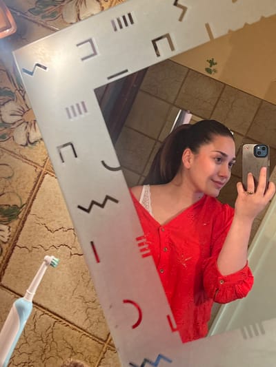 The image displays a mirror selfie taken within an interior space, likely a bathroom, located in Ancenis, France. The central subject is a female individual reflected in a rectangular mirror, which has a frosted border adorned with various geometric cut-out shapes including lines, squares, zigzags, and arcs. The individual possesses dark hair pulled back into a ponytail. She is wearing a red, long-sleeved, collared shirt with embroidered or eyelet details, unbuttoned to partially reveal a white lace undergarment. Her left hand holds a dark-colored smartphone, likely an iPhone due to the triple camera module visible, which is being used to capture the image. A gold-colored ring is present on her left ring finger. Her head is slightly turned to her left, and she exhibits a subtle smile. In the lower-left corner of the foreground, outside the mirror's reflection, a light blue and white electric toothbrush with a white brush head rests on a beige, patterned tiled surface. The visible background, both within and outside the mirror's reflection, consists of light-colored, possibly ceramic, tiles featuring a distressed texture and some floral or organic patterns.