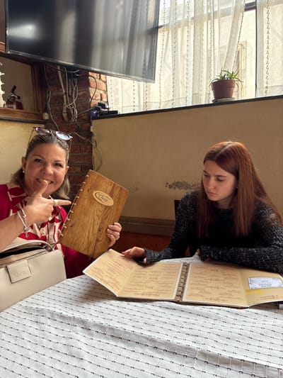 Two women are seated at a table covered with a white tablecloth with a grid pattern. The woman closest to the viewer is smiling and holding a wooden menu open with her right hand. She is wearing sunglasses on her head, a red and white patterned shirt, and has a light-colored purse next to her. The second woman, with red hair, is looking down at another menu. She is wearing a black sweater. Both menus are open, showing text in Spanish. Behind the women, there is a television mounted on a brick wall with exposed wires. To the left of the TV, a window shows a potted plant and partially visible cityscape through sheer curtains. The setting appears to be a restaurant. The wooden menu has a logo of "La Asoma Restaurante Bar" indicating the location is in Tepeji de Ocampo, Mexico.