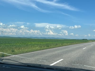The image captures a view from a vehicle, looking out at a vast agricultural landscape under a partly cloudy blue sky. The foreground shows the dashboard and windshield of the vehicle, with wipers visible. A grey asphalt road with white markings stretches into the distance, bordered by a vibrant green field. The field appears to be planted with crops, possibly corn or soybeans, extending to a line of distant hills. The sky is a brilliant blue, dotted with fluffy white cumulus clouds and streaky cirrus clouds. The location is identified as Shoshino, Russia, suggesting a rural or semi-rural setting with extensive farmland. There are no people or animals visible in the scene. The overall impression is one of a bright, sunny day in the countryside.
