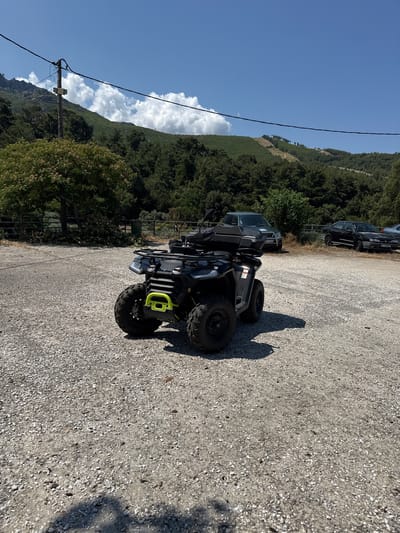 A black ATV is parked on a gravel surface in Rachoni, Greece. The ATV is facing slightly to the left, with its front wheels visible. Behind the ATV, several vehicles are parked: a dark-colored SUV, a black sedan, and partially visible, a silver car. A fence runs horizontally across the middle ground, with trees and vegetation on the other side. In the background, a lush green, tree-covered hillside rises under a bright blue sky with scattered white clouds. A utility pole with wires is visible on the left side of the image. The scene appears to be an outdoor parking area in a natural, possibly rural, setting.