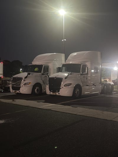 Two white semi-trailer trucks are parked in a lot at night, illuminated by a bright overhead light. The truck on the left is a 2020 International LT, with the logo "ASL Transportation" visible on its side. The truck on the right is a newer model, also white with the "ASL Transportation" logo. Both trucks are clean and appear to be in good condition. The parking lot is paved and appears to be empty except for the trucks. In the background, there are some trees and buildings, as well as other vehicles. The sky is dark and clear, with no visible stars. The overall impression is one of a quiet, industrial area at rest. The image is undated and no specific location is provided beyond "New Britain, United States of America".