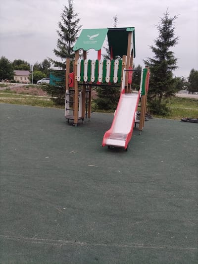 An outdoor shot captures a playground with a slide, stairs, and a green roof structure. The playground is situated on a dark green rubberized surface, and trees are visible in the background, along with a building and a car. The sky is overcast. The playground is located in Nizhnyaya Makhtama, Russia.
