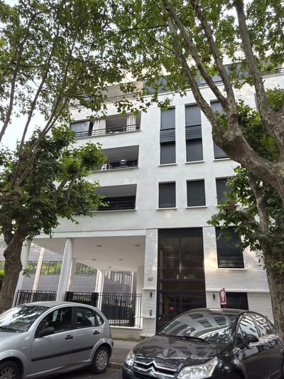 The image depicts a street scene in Maisons-Alfort, France, focusing on a modern white apartment building with balconies and several windows. The building is partially obscured by the branches of mature trees with lush green leaves. In the foreground, two cars are parked on the street. The car on the left is a silver hatchback, and the car on the right is a dark-colored hatchback. A black metal fence runs along the front of the apartment building's ground level, and a pedestrian entrance with large glass windows is visible. The building has a prominent number "167" displayed next to the entrance. The sky is overcast.