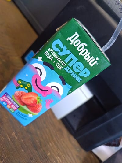 An overhead close-up shot captures a juice box with a straw. The juice box is primarily blue with a green top and features a cartoonish face with large eyes and a pink tongue. Strawberries and a video game controller are depicted on the blue part of the box. The Russian text on the box translates to "Dobry Super Drink". The box is angled on a wooden surface, suggesting it is on a table. The background is blurred, but appears to be a dark electronic device. The scene is static and no people are present. The location in Knevichi, Russia is not discernible from the visual information provided.