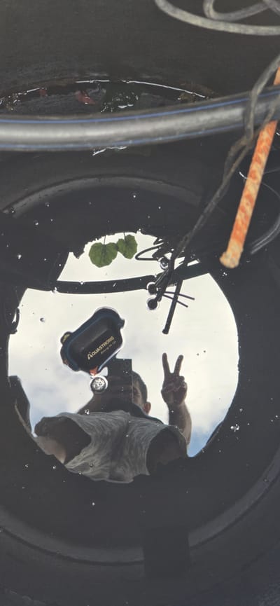 The image shows a reflection in water within a dark, cylindrical structure, possibly a manhole or a large pipe opening, located in Русская Деревня-2 Кп, Russia. The reflection reveals a person holding a smartphone, taking a selfie, and making a peace sign with their right hand. The person is wearing a light-colored shirt. Floating in the water are a few green leaves and an object that appears to be a waterproof phone case with "AQUASTRONG" written on it. The sky and clouds are visible in the reflection, indicating the photo was taken outdoors during the day. Cables and a rusty metal rod are also visible in the structure above the water.