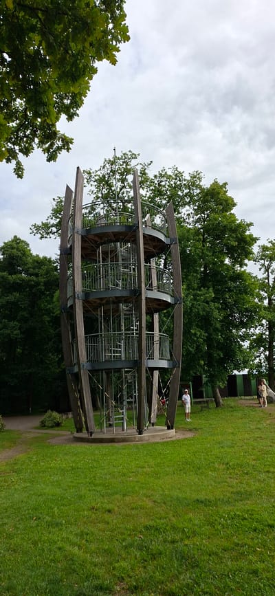 The image shows a multi-level spiral observation tower made of wood and metal, located in Valgeranna, Estonia. The tower has several circular platforms connected by a central spiral staircase. It is surrounded by lush green grass and mature trees. In the background, there are some people and a dark green building. The sky is overcast with clouds. The tower appears to be a tourist attraction or a recreational facility within a park-like setting.