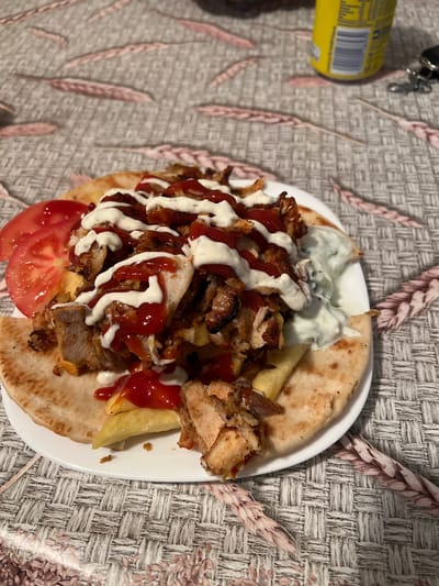 The image shows a gyros dish served on a white plate. The gyros consists of pita bread, sliced gyro meat, tomato slices, fries, a white sauce (possibly tzatziki), and ketchup. The plate is placed on a textured surface with a pattern resembling woven fabric with pale wheat motifs. In the upper left corner of the image, there is part of a yellow can and keys. The location is likely not Dushevo, Bulgaria as this is not discernible from the image.