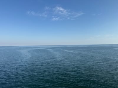 A serene, wide-angle shot captures a tranquil seascape under a clear blue sky. The horizon line is sharply defined, separating the deep blue, gently rippled ocean from the pale blue sky above. Wispy white clouds are scattered across the upper portion of the sky, adding subtle texture. The ocean surface is consistent, with small, regular waves creating a mesmerizing pattern of light and shadow. The scene is devoid of any human presence, vessels, or landforms, suggesting an open expanse of water. The lighting indicates it is daytime, likely with the sun positioned high in the sky. The location is described as Unknown, Lithuania, but the image itself provides no specific geographical markers to confirm this.