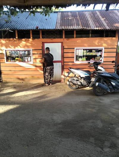 A man stands at the open doorway of a small orange-painted shop in Sagoe, Indonesia. The shop has a corrugated metal roof with two display windows on either side of the door. The windows contain items for sale, including food and drinks. Two motorcycles are parked to the right of the shop's entrance. The ground in front of the shop is dirt and gravel. The man is wearing a dark t-shirt, camouflage pants, and sandals. He has short dark hair. The lighting indicates it is daytime.