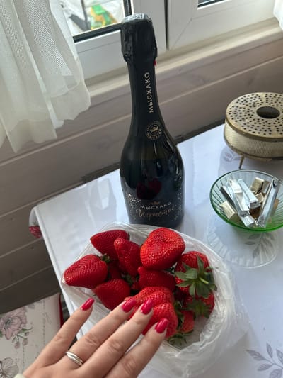 On a table in Bashur, Russia, a still life is arranged. A bottle of sparkling wine labeled "Myskhako Igristoe" lies next to a clear plastic container filled with strawberries. A woman's hand with red nail polish is reaching into the strawberries. A small green glass bowl containing foil-wrapped candies is positioned next to a cylindrical metal object with a perforated top. A white sheer curtain is visible in the background near a window. The table is white.