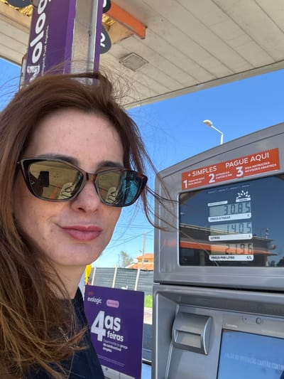 A female individual with reddish-brown hair and freckles on her face is partially visible, positioned in front of a fuel dispenser at a gas station in Alcochete, Portugal. She wears dark, cat-eye style sunglasses, which reflect a smartphone in the left lens.
The prominent silver-grey fuel dispenser on the right features an upper digital display showing "PREÇO A PAGAR 30.85 €," "LITROS 14.05 L," and "PREÇO POR LITRO 2.196 €/L." Above these figures, instructions in Portuguese read: "PAGUE AQUI," "1 SIMPLES TOQUE NO ECRÃ PARA INICIAR," "2 INTRODUZA CARTÃO E PIN," and "3 SIGA INSTRUÇÕES NO VISOR E ABASTEÇA." Below the main display, a second screen, labeled "Petroféc," shows a partial message: "FALHA NA OPERAÇÃO CARTÃO CONTIN..." (Card operation failed...). A card slot is visible beneath the upper screen.
The background features a clear blue sky with a visible street light. A white gas station canopy is partially visible overhead. To the left, a purple vertical sign with "golo Intelligent Fuel Agents" and a partial number "2" is present. Another purple sign below reads "evologic" and "4as feiras Experimente o preço do combustível simples." Distant structures, including a building with an orange roof and a grey fence, are discernible in the far background.