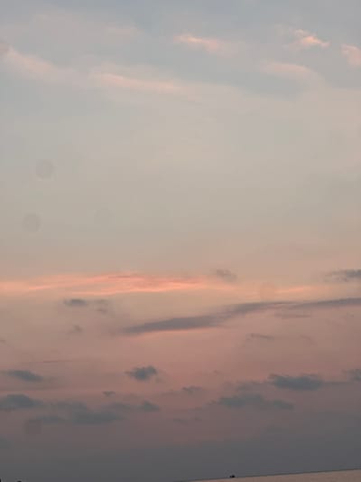The scene captures a panoramic view of the sky and a body of water in Preah Sihanouk, Cambodia, characteristic of either dawn or dusk. The sky, occupying the majority of the frame, displays a color gradient from a light blue-grey at the top, transitioning downwards through faint pink and orange hues, to darker purplish-pink tones at the lower edge. Scattered cloud formations are visible, ranging from wispy cirrus-like streaks in the upper sky to more defined, broken cumulus-like clouds with varying shades of grey and pink towards the middle and lower sky. A distinct, calm body of water, consistent with the Gulf of Thailand, is visible at the very bottom of the image, separated from the sky by a clear, horizontal horizon line. A single, small, dark-hulled vessel is present as a silhouette on the water, located slightly to the right of the frame's center in the lower quadrant. Two faint, circular, semi-transparent smudges are observable in the upper-left quadrant of the sky.