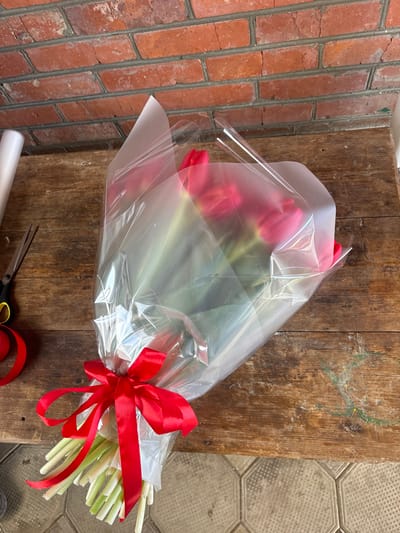 An overhead view reveals a freshly arranged bouquet of red tulips, wrapped in clear plastic film and white paper, secured with a red satin bow, positioned on a weathered wooden surface in Trudobelikovskiy, Russia. The bouquet consists of multiple red tulips with visible green stems, cut bluntly at the bottom. To the left of the bouquet, a roll of white translucent wrapping material stands vertically, alongside a pair of black-handled scissors with yellow finger loops, and a coil of red satin ribbon. The wooden surface exhibits visible grain and some green/blue stains. A red brick wall forms the immediate background, while a floor composed of light-colored octagonal or hexagonal tiles with dark grout lines is partially visible in the lower foreground. No individuals are present in the scene.
