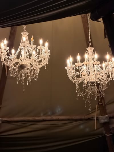 The image depicts two ornate chandeliers hanging in what appears to be a tent or rustic indoor space. The chandeliers are made of white metal and adorned with numerous crystal pendants and candle-shaped lights, all illuminated. The background is a muted, tan fabric. Wooden beams and dark fabric elements are visible, suggesting a temporary structure. There are no people or animals in the image. The scene is dimly lit, with the chandeliers being the primary light source. The location is identified as Cavriglia, Italy.
