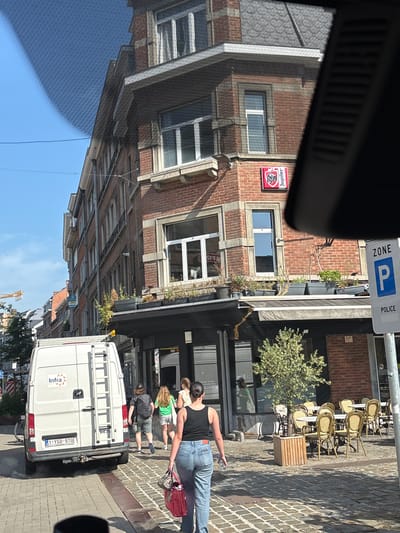 The image depicts a street scene in Namur, Belgium. A white van with a license plate reading "1-YSD-570" and a ladder on its roof is parked on the left side of the cobblestone street. To the right of the van, several people are walking. One person is wearing a black tank top and blue jeans, carrying a red bag. Further to the right, an outdoor seating area with tables and chairs is set up in front of a brick building. A sign on the building displays the "Jupiler" logo. A blue and white "ZONE P POLICE" sign is visible on a pole on the far right. The buildings are made of brick and have multiple windows. The sky is clear and blue.