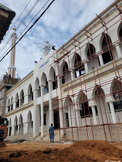 The image depicts a large, white mosque building under construction or renovation, viewed from a slightly low angle. The main structure is adorned with extensive red metal scaffolding, covering its facade and parts of the upper sections.

The building features a distinctive architectural style with multiple arched openings and decorative elements. A prominent horizontal band of intricate calligraphy, rendered in gold or yellow on a dark blue background and framed by lighter ornamental patterns, runs along the upper part of the main facade. Below this band, a series of white arched recesses or windows are visible.

To the left of the main building, a tall, slender minaret rises, featuring alternating white and gold vertical stripes and topped with a golden finial. Another minaret-like structure or dome area on the building itself is also covered in scaffolding, with what appear to be workers present high up.

On the right side of the image, the foreground is dominated by rough, reddish-brown earth or dirt, suggesting recent excavation or a construction site. Next to the building, on a seemingly paved area or steps, a man in a light blue traditional garment (possibly a thobe) stands facing the building. Partial views of an orange construction vehicle are visible at the very bottom, near the dirt and the building.

The sky above is overcast with patches of blue visible through white clouds. Several dark power lines traverse the upper left portion of the frame.

A blue sign with white text is visible vertically on the building's facade, clearly reading "JAMII MASJID". The overall scene suggests ongoing work on a mosque during daylight hours, under cloudy conditions.