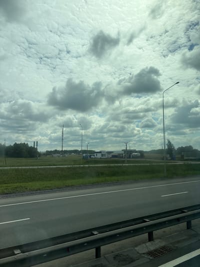 The image captures a roadside scene on a cloudy day, likely in or near Poisevo, Russia. A multi-lane asphalt road runs horizontally across the foreground, separated by white lines. A metal guardrail is positioned between the road and a grassy embankment. Beyond the embankment, a flat, open field is visible, dotted with a few scattered buildings and structures.

Prominent features in the background include several tall communication towers, some with visible antennas. There appears to be a small cluster of buildings that could be a gas station or service area, with a white structure bearing what looks like Cyrillic text and a smaller building with a blue roof. A few trees line the horizon, and a single streetlamp stands near the right side of the frame, with another smaller one visible below it. The sky is filled with a dense layer of cumulus clouds, obscuring much of the blue sky. The overall impression is one of a rural or semi-urban landscape along a transportation route. There are no people or animals visible in the image.