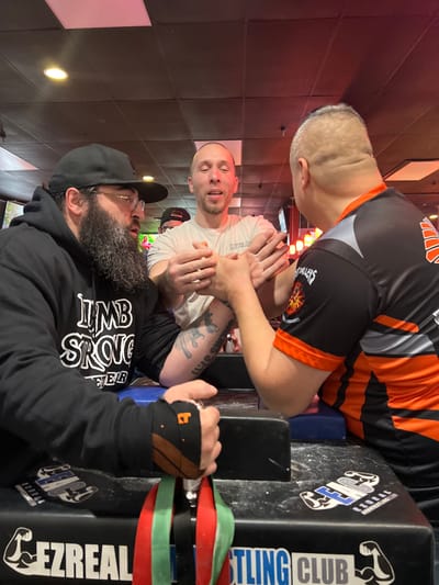 Three men are arm wrestling. Two men are positioned on either side of the arm wrestling table, clasping hands in preparation to begin. One man wears an orange and black jersey while the other wears a white t-shirt. A third man with a beard, wearing a black hoodie and cap, looks on. The arm wrestling table is black with "Wrestling Club" and "EZREAL" written on it. The background includes a black and white checkered wall and some red decorative elements.