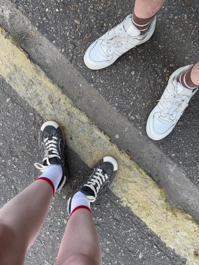 Three pairs of legs and feet wearing sneakers stand on asphalt pavement with a yellow painted line. Two pairs of feet on the left wear white Adidas sneakers and brown socks with white stripes. The pair of feet on the right wear dark gray and white sneakers with white socks that have red stripes. The location and city of Balezino, Russia, cannot be confirmed without further evidence.