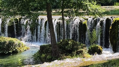 The image shows a cascading waterfall in a natural setting, likely in or near Vrbica, Bosnia and Herzegovina. Lush green vegetation, including trees with dense foliage, surrounds the waterfall. The water flows over moss-covered rocks, creating a frothy, white curtain. The stream in the foreground is clear and reflects the green of the surrounding plant life. There are no people visible in the image, and the scene depicts a tranquil, natural environment. The lighting suggests it is daytime, with sunlight filtering through the trees.