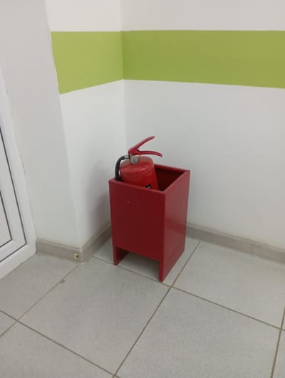 A red fire extinguisher is placed inside a red metal stand in the corner of a room. The room has white walls with a horizontal green stripe across the upper portion. The floor is tiled with light-colored square tiles. To the left of the fire extinguisher, a portion of a white door frame is visible. The scene appears to be an interior public space, likely in a building in Alğa, Kazakhstan. There are no people or any overt actions occurring in the image.