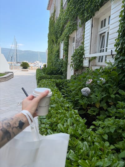 The image shows a person holding a disposable coffee cup and a white paper bag. The person's arm has tattoos. The background features a building covered in ivy, white shutters on windows, and lush green foliage with some hydrangeas. Beyond the foliage, a paved walkway leads towards a view of a bay with sailboats and mountains in the distance. The sky is clear and blue, suggesting a sunny day. A small palm-like plant is visible on the walkway. The scene appears to be outdoors, likely in a coastal or harbor area. The text on the coffee cup is partially visible and appears to say "BAGUETTES". The overall mood is relaxed and leisurely, perhaps a person enjoying a morning coffee while on vacation or a stroll. The image is oriented vertically, with the scene appearing as if viewed from a slightly elevated position looking down.