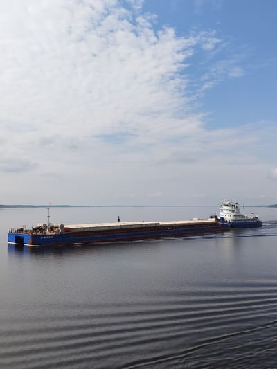 A high-angle, full shot depicts a large cargo barge traveling on a body of water under a partially cloudy sky. The barge is blue and has a brown rust-colored waterline. It is loaded with rectangular cargo. A white tugboat is connected to the rear of the barge. The wake of the vessel creates ripples on the water's surface. A distant shoreline is visible on the horizon. Based on visual observation and publicly available information, it can be inferred the photo was taken in the vicinity of Глебово, Russia.