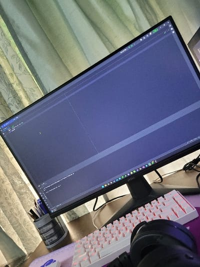 A tilted view of a desk setup. A large monitor dominates the frame, displaying code or a similar application on its screen. To the right, a white keyboard with red accents is visible, alongside a pair of black headphones. A small desk organizer with pens and pencils sits near the monitor's base. The scene includes a textured desk surface and a glimpse of patterned curtains or blinds in the background.