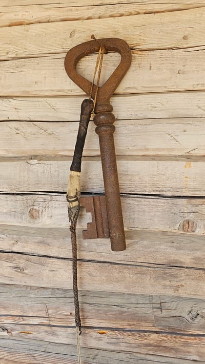 A rusty, oversized, antique iron key is displayed on a weathered wooden wall. The key has a large, heart-shaped bow and a long, cylindrical shaft with a multi-tiered bit. A thin, worn leather strap hangs from the key's bow, attaching it to the wall and also connecting to a braided leather whip or lash that dangles below. The whip has a fur-covered section near its attachment point to the key. The wall is constructed of horizontal, rough-hewn wooden planks with visible grain, knots, and signs of age. The lighting is natural and appears to be daylight, casting subtle shadows. The scene suggests an outdoor or rustic setting. The location is the courtyard of a historic homestead in Tylinava, Lithuania.