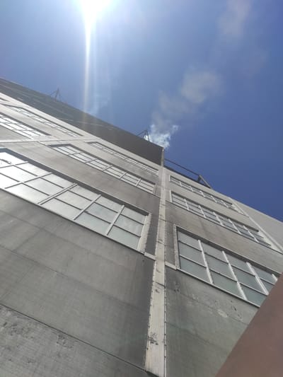 The image is an upward-angle shot of a tall, industrial-looking building with a grey, textured facade and numerous large, multi-paned windows. The building is seen from a low perspective, emphasizing its height against a bright blue sky with some wispy white clouds. The sun is very bright and positioned in the upper left corner, creating lens flare and washing out parts of the sky. A thin plume of white smoke or steam is visible rising from the top of the building against the sky. The building appears to be made of concrete panels, with vertical seams and some signs of wear and tear on the facade. The windows are framed in a light color, possibly white or silver, and reflect the bright sky. The scene appears to be in an urban or industrial setting. There are no people visible in the image. The building's architectural style suggests it could be a factory, warehouse, or older commercial building. The location of Kyzyl, Kazakhstan is not explicitly shown or implied by any visual elements in the image itself, but is provided as context.