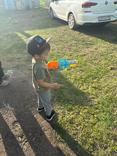 In a rural outdoor setting, likely a backyard in Чуй-Атасево, Russia, a young child, wearing a black baseball cap with Star Wars motifs, a green t-shirt with a dinosaur print, and grey sweatpants, stands on a gravel path, holding a blue and orange toy water gun. The child appears to be in the process of playing, with a stream of water visibly spraying from the gun towards the right side of the frame. In the background, a white Volkswagen sedan is parked on a grassy area. The environment is characterized by a weathered wooden structure to the left, a patch of dirt path, and a sun-drenched lawn with scattered toys, suggesting a residential area. The lighting indicates it is daytime, with long shadows cast by the sun.