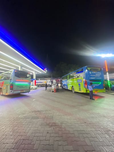 At a brightly lit gas station in Kajheri, India, a fleet of buses and trucks are parked under a covered canopy illuminated by white and blue LED lights. The ground is paved with interlocking bricks.

On the left, a white and green bus with "TRAVEL AP" written on its rear is parked. The bus's taillights are illuminated with a red and green pattern.

In the center, a white car is parked, and further back, a fuel tanker truck with "POWERES" written on it is also visible.

On the right, a blue and green IntrCity smartbus is parked. A man in a blue uniform, likely an attendant, stands near the rear of the IntrCity bus, holding a device. The bus features "IntrCity smartbus" branding and contact information. Green and orange lights are visible to the right of the bus. The scene is captured at night.