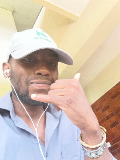 An adult male, appearing of African descent, is depicted in a close-up, selfie-style photograph. He wears a light grey baseball cap with a green "NEDBANK" logo on the front, a light blue button-up shirt unbuttoned at the collar, and white wired in-ear headphones. His right hand is raised to chest level, performing a "shaka" gesture with the thumb and pinky finger extended. On his right wrist, he wears a coiled gold-toned bangle above a silver-toned segmented metal bracelet. He looks directly at the camera with a slight smile. The background features light-colored overhead architectural elements to the upper left and a reddish-brown brick wall to the right. The specific location within Chinhoyi, Zimbabwe, is not visually identifiable from the depicted scene.