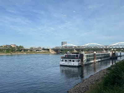 The image depicts a wide river, identified as the Rhine, in Ludwigshafen am Rhein, Germany, under a clear, light blue sky with wispy clouds. In the midground, a large white river cruise ship, named "EMILY BRONTE", is docked on the right bank. Its stern is visible, displaying "BASEL" and "CH" with a Swiss flag, indicating its homeport. The ship has multiple decks with large windows and is connected to the shore by a gangway. The river's surface is dark blue with noticeable ripples, reflecting the sky and surrounding light.
On the immediate right riverbank, a rocky embankment (riprap) extends along the water's edge, partially covered by green bushes and foliage. In the background, multiple bridges span the river. The most prominent bridge features white, arched truss structures with grey supports, extending across the full width of the river. Another darker, lower bridge, possibly a railway bridge, is visible beneath and to the left of the white-arched bridge. Further back on the left, additional elevated road or rail structures are present.
The far bank of the river showcases an urban landscape. On the left, a cluster of multi-story, light brown and reddish buildings with pitched roofs is visible. Towards the center-right of the far bank, a tall, rectangular skyscraper with a dark, possibly glass, facade rises prominently above the other structures. Various other urban buildings and infrastructure, including power lines or catenary structures, are scattered along the far bank, with patches of green trees and vegetation interspersed within the urban fabric. No people are distinctly visible.