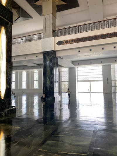 The image depicts the interior of a modern mosque located in Aluekambuk, Indonesia. The spacious hall features a highly polished dark green marble floor that reflects the ambient light. Several large, white marble columns with intricate geometric carvings support the high ceiling, which is adorned with dark and light patterned panels. A second-level balcony with ornate black railings and decorative Islamic calligraphy is visible in the background. Large windows with patterned screens let in diffused natural light, creating a bright and airy atmosphere. In the foreground, a young child is seen walking across the vast floor. To the left, a section of a tall, illuminated decorative element with a pointed arch design is visible. The overall impression is one of grandeur and tranquility.