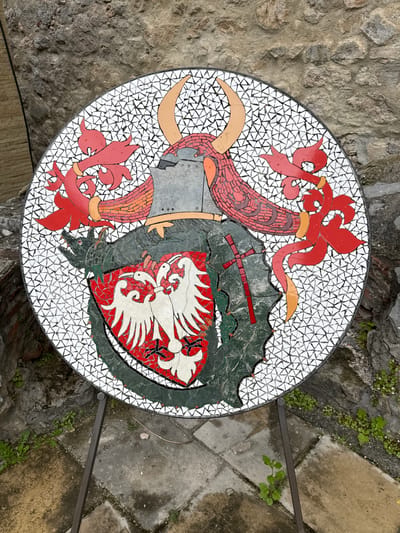 The image depicts a mosaic table top outdoors in Despotovac Municipality, Serbia. The mosaic features a coat of arms with a helm, dragon, and shield containing white birds on a red background. The background of the mosaic is white with triangular tesserae. The table is set against a backdrop of stone walls and paved surfaces, with small plants growing nearby.