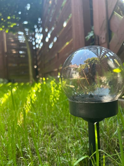 This is a close-up, eye-level, outdoor photograph of a solar garden light in a grassy yard, likely in Środa Wielkopolska, Poland. The solar light, a clear glass orb with internal copper wire filaments, sits on a black base and stake, inserted into lush green grass. The orb reflects the surrounding environment, showing a wooden fence, trees, and a building with a peaked roof and windows. Sunlight filters through the leaves of a tree, creating a bokeh effect of bright, blurry circles in the background, illuminating the grass with dappled light. The fence, made of vertical wooden planks, runs horizontally across the mid-ground, creating a textured backdrop. The overall scene suggests a tranquil, daytime setting.