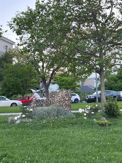 The image depicts a street scene in Neberdzhaevskaya, Russia, with a focus on a residential area. In the foreground, a well-maintained lawn with small white flowers is visible. A decorative stone structure, possibly a planter or garden feature, stands to the right of the frame. A woman on a bicycle, wearing a white cap and a white t-shirt, is seen riding past. Behind her, a red car is parked. Further in the background, several other vehicles, including a dark-colored SUV and a white sedan, are parked along the street. Trees with lush green foliage are interspersed throughout the scene, providing shade and a natural element. Residential buildings, with their characteristic architecture, are visible in the background, suggesting an urban or suburban setting. The sky is overcast, indicating a cloudy day. The overall impression is one of a quiet, residential street, possibly in a town or city, with everyday life unfolding.