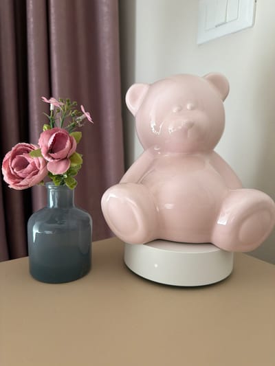 A light pink, glossy ceramic teddy bear figurine sits on a white circular base to the right of a dark gray glass vase holding a small bouquet of artificial pink flowers. The scene takes place indoors, on a light tan surface, with a textured purple curtain in the background on the left and a white wall with a light switch on the right. The lighting is soft and natural. The teddy bear is in a seated position, facing forward with its paws resting on its knees. The flowers in the vase are a mixture of pink roses and smaller pink blossoms with green leaves and stems. The overall aesthetic is decorative and appears to be part of a home or shop interior in Spinea, Italy.