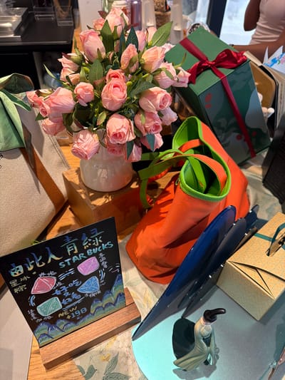 The image captures a still life arrangement on a table, possibly in a Starbucks store in Jianye District, China, given the prominent "Starbucks" branding and Chinese characters. The central focus is a bouquet of pink artificial roses in a white vase, resting on a wooden block. To the left, a beige canvas tote bag with a brown strap is visible.

In front of the flowers, a small blackboard sign displays colorful drawings of stylized mountains, what appear to be ice candies in pink, blue, and green, and Chinese text which includes "STARBUCKS" and "NEW ¥198".

To the right of the flowers, an orange tote bag with a green lining and straps is prominently displayed, partially obscuring a tall green gift box tied with a red ribbon. Further to the right, a light yellow gift box with a geometric pattern is visible. A small, delicate figurine of a person in a green robe and black hat is positioned in the foreground, casting a shadow. The overall scene is well-lit, suggesting an indoor environment with natural light.