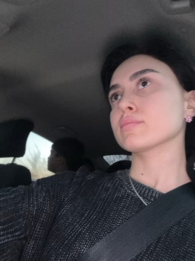 The image depicts the interior of a passenger vehicle, purportedly in Kizil-ajlo, Georgia. In the foreground, a female with dark hair and a light complexion is visible from the chest up, positioned towards the upper right of the frame. She wears a dark gray, textured knit sweater, a thin silver chain necklace, and a black seatbelt diagonally across her torso. A pink, flower-shaped earring is present on her right earlobe. Her head is tilted slightly upwards and to her right, with her gaze directed in that direction. In the mid-ground, behind the primary subject, the back of another individual's head with short dark hair is partially visible, appearing to look out the left side window of the vehicle. The vehicle's gray ceiling is visible in the upper portion of the frame. Blurred natural scenery is visible through the vehicle's window in the background.