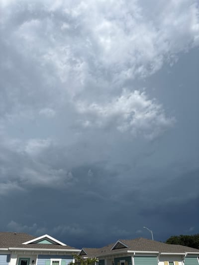 An overcast sky looms above two houses in Sarasota, Florida. The clouds are dark and heavy, suggesting an impending storm, with lighter clouds scattered across the upper portion of the frame. The houses, painted in light blue and white with light teal accents, have gray shingled roofs. A streetlight is visible above the right house. The image is captured from a low angle, emphasizing the dramatic sky. There are no people or animals visible in the image.