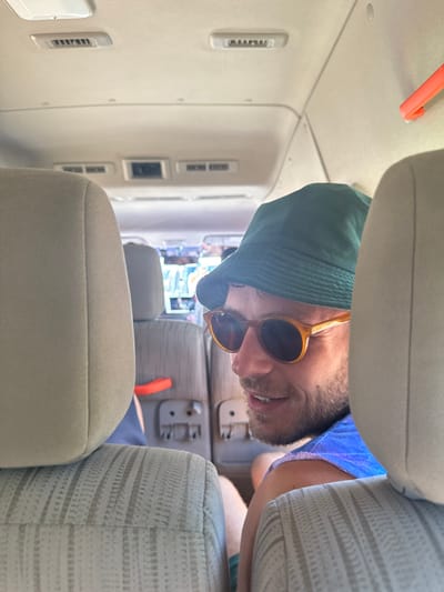 The image depicts the interior of a light-colored passenger vehicle, likely a van or shuttle. An adult male is featured prominently in the mid-right of the frame, partially obscured by the foreground seatback. He is wearing a dark green bucket hat and light brown-framed sunglasses, and has facial stubble. He is smiling faintly, looking towards the left. He wears a blue sleeveless top. The vehicle's interior features light beige, patterned fabric seats, with two seatbacks visible in the foreground. Overhead, white ceiling panels contain air vents and light fixtures. An orange plastic handle is visible on the side panel behind the left foreground seat. The background, visible through the vehicle's front, is blurry and shows indistinct colorful shapes, suggesting an outdoor environment. The image does not contain specific visual identifiers to confirm the exact location within Falmouth, Jamaica.