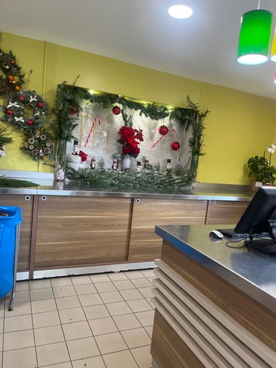 The scene depicts the interior of a commercial establishment in Aiguilhe, France, decorated with a Christmas or winter theme. The walls are painted a light yellow-green, complementing a light-colored tiled floor. Overhead, recessed circular lights and green cylindrical pendant lights illuminate the area.

On the left wall, multiple evergreen wreaths, adorned with red berries, white flocking, pinecones, and dried orange slices, are displayed alongside evergreen garlands. Spanning the central background is a prominent display set against a light-colored panel and framed by evergreen garlands with small white lights. This display features several red Christmas baubles, large red-and-white candy cane decorations, and a central arrangement of red poinsettia flowers in a grey cylindrical pot. Below this central display, miniature snow-dusted evergreen trees and small decorative figures, resembling nutcrackers, are arranged along a surface. A tall glass vase containing green foliage and small pink or white decorative items stands to the left of this arrangement.

A long counter with a stainless steel top and wooden cabinets with sliding doors extends across the mid-ground. To the far left, a blue plastic waste bin on a metal frame is visible. In the foreground on the right, a point-of-sale or service counter with a stainless steel top and wooden side panels displays a black computer monitor, a white computer mouse, and a black keyboard. In the far right background, a potted plant with large green leaves and white orchid flowers is present. No people are visible within the scene.