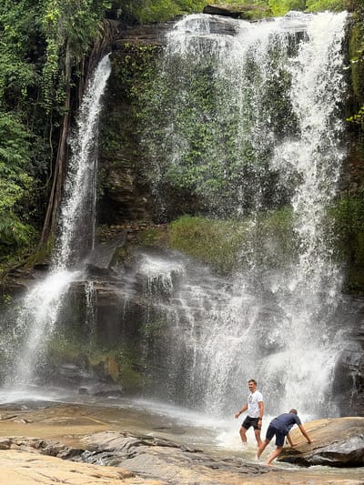 Two men are at the base of a large waterfall, likely located in Ban Huai Takhrai, Thailand. The waterfall cascades down a rocky cliff face covered in lush green vegetation. The water is a white, frothy spray, creating mist that obscures some of the background. The men are positioned in the shallow water at the bottom of the falls. One man, wearing a white t-shirt and black shorts, stands facing the waterfall with his arms slightly out to his sides. The other man, wearing a blue t-shirt and dark shorts, is crouched down, with his hands on a large, wet rock, appearing to be climbing or steadying himself. The ground surrounding the pool at the base of the waterfall is covered in rocks of varying sizes, some smooth and wet, others rougher and drier. The overall scene depicts a natural, adventurous setting with the powerful force of the waterfall as the dominant element.
